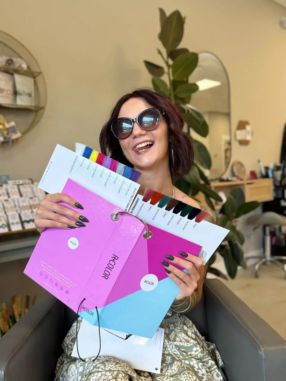 Em laughing in a salon chair, sunglasses on, holding up a ring of bright hair color swatches.